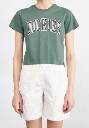 Kobieta w zielonym, krótko skrojonym t-shircie marki Dickies i białych, wysokich szortach cargo, stoi na jednolitym tle.