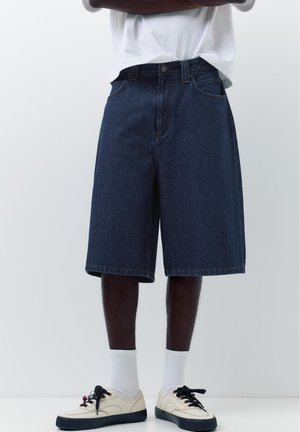 Osoba nosząca ciemnoniebieskie dżinsowe spodenki do kolan, białe skarpetki oraz kremowe trampki z czarnymi sznurowadłami, stojąca z założonymi ramionami.