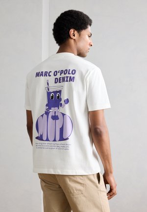 Άνδρας που φοράει λευκό μπλουζάκι με μοβ καρτούν ποτό και το κείμενο «MARC O'POLO DENIM» στην πλάτη, συνδυασμένο με μπεζ παντελόνι.