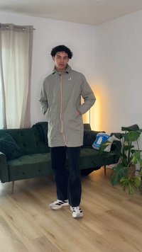 Joven con chaqueta gris clara y pantalones negros, de pie en una sala de estar moderna con sofá verde, plantas y suelo de madera.