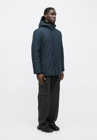 Muž stojaci, oblečený v námornícky modrej kapucňovej zimnej bunde, čiernych cargo nohaviciach a čiernych teniskách na jednofarebnom svetlom pozadí.