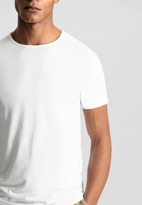 Maglietta bianca a maniche corte realizzata in tessuto liscio, con scollo tondo e dettagli di cucitura minimal. Indossata con pantaloni beige.