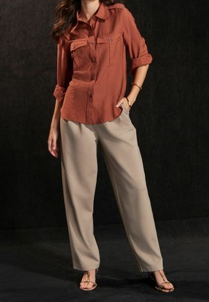 Rust-colored button-up shirt with two front pockets, paired with beige wide-leg pants and strappy sandals on a dark background.