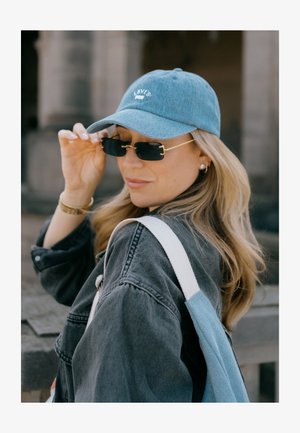 Denim kapa v svetlo modri barvi z belim logotipom, pravokotna črna sončna očala z zlatimi detajli, siva denim jakna in torba preko rame z modrimi in belimi trakovi.