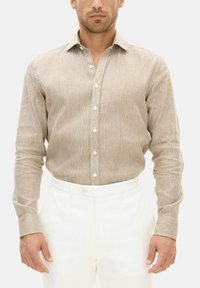 Camicia beige a maniche lunghe con sottili righe verticali e colletto classico. Presenta bottoni bianchi e una texture liscia. Indossata con pantaloni bianchi.