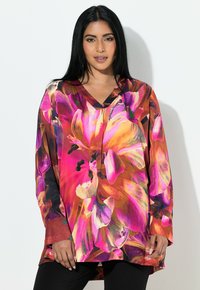 Kvinde med langt sort hår iført en farverig, stor blomstret bluse med lyserøde, orange og lilla kronblade, poserer mod en hvid baggrund.