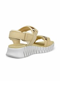 Sandalia de un beige claro con un empeine sintético texturizado, doble tira y acentos metálicos. Suela blanca gruesa con un patrón ondulado para mayor agarre.