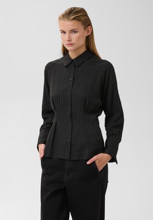 Jeune femme aux cheveux blonds raides porte une chemise noire cintrée à boutons et un pantalon noir, debout les mains dans les poches sur un fond uni.