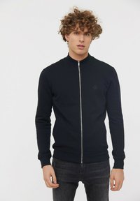 Veste zippée noire avec col montant, manches et ourlet côtelés, fabriquée en tissu lisse, avec un petit logo sur la poitrine.