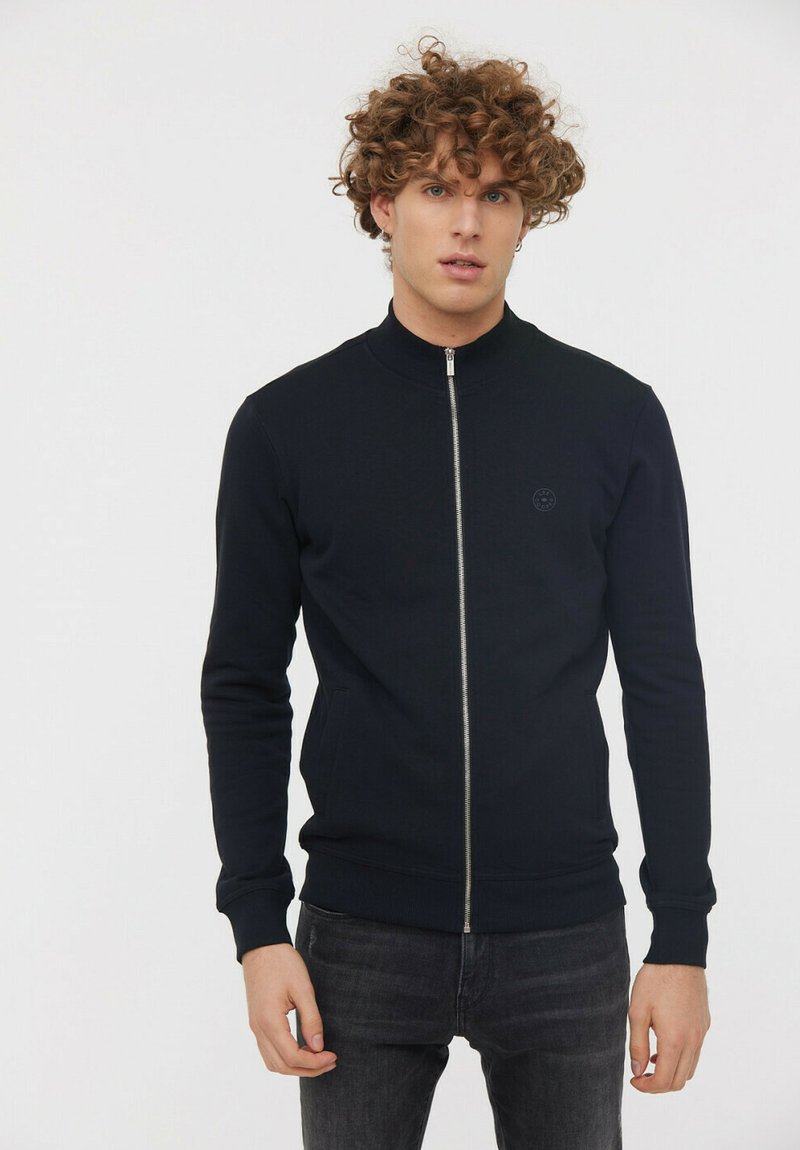 Veste zippée noire avec col montant, manches et ourlet côtelés, fabriquée en tissu lisse, avec un petit logo sur la poitrine.