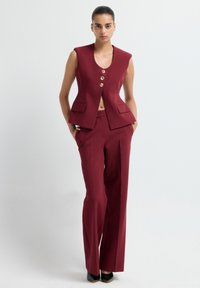 Gilet sans manches bordeaux avec un décolleté en V profond, quatre boutons dorés et deux poches avant, associé à un pantalon large assorti.