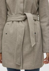 Beige villakangastakki, jossa on solmittava vyö, kaksi etutaskua tummilla napeilla, teksturoitu kangas ja jäsennelty muotoilu.