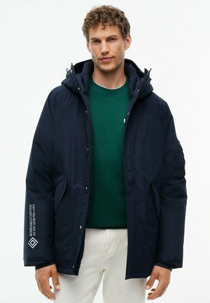 Mann trägt eine dunkelblaue Kapuzenjacke mit weißem Text auf dem Ärmel, grünen Pullover und weiße Hose, steht vor einem einfarbigen Hintergrund.