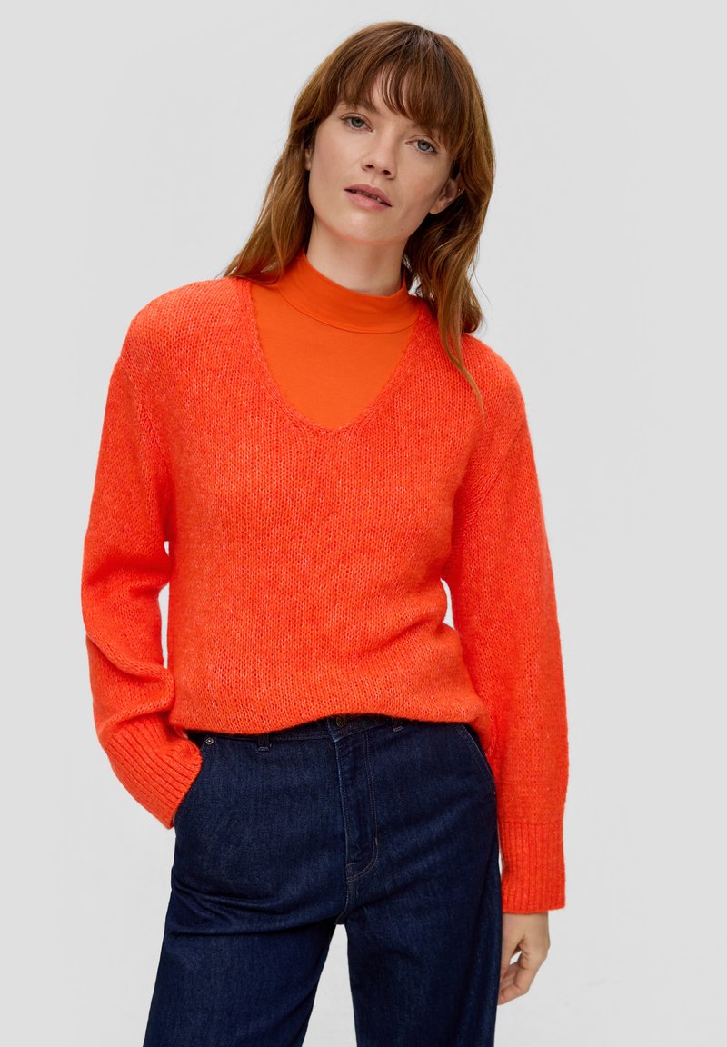 Oranje gebreide trui met een diepe V-hals, met lange mouwen en een losse pasvorm. Gecombineerd met een feloranje coltrui eronder.