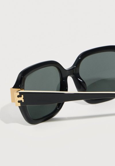 Lunettes de soleil carrées oversize noires avec des verres foncés et un détail en métal doré sur les branches, sur un fond clair.