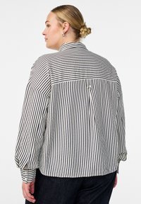 Küps blondide juustega naine, kel on juuksed kuklapunsus, kannab laia musta-valge vertikaalse triibuga särki ja tumedaid pükse, seistes vasakul valgel taustal.