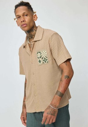 Jeune homme avec des tatouages au cou et aux bras portant une chemise beige texturée à manches courtes avec un patch brodé et un pantalon sombre, se tenant devant un fond uni.