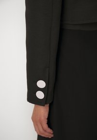 Gros plan d'une manche de veste noire texturée avec deux gros boutons blancs près du poignet et une main détendue le long du corps.