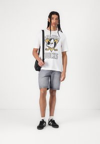 T-shirt en coton blanc avec un graphique "ANAHEIM DUCKS", shorts en denim gris, baskets noires et un petit sac bandoulière noir.