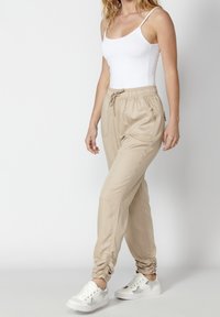 Pantalones jogger beige con cintura elástica y puños en los tobillos, que cuentan con bolsillos laterales, combinados con una blusa sin mangas blanca y zapatillas blancas.