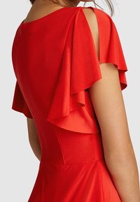 Robe rouge avec des manches volantes, un corsage ajusté et une texture lisse. Le dos présente un détail de fermeture éclair discret.