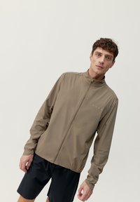 Ljusbrun zip-up jacka i mjukt tyg; har hög krage, långa ärmar och diskret logotypdetalj på bröstet.