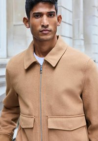 Chaqueta de mezcla de lana color marrón claro con cierre de cremallera en el frente, cuello con puntas y dos bolsillos de parche en el pecho. Textura suave y ajuste entallado.