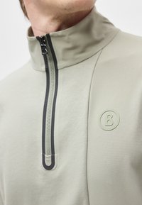 Κοντινό πλάνο ενός ατόμου που φοράει ανοικτό γκρι τοπ με φερμουάρ με μαύρη περίμετρο και κυκλικό λογότυπο στο στήθος.