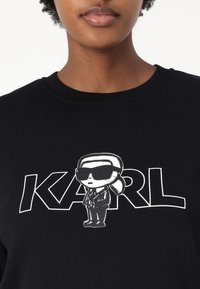 Črna majica s kapuco, na kateri je risanka s karakterjem v sončnih očalih in obleki, z besedilom "KARL" v bold belem fontu. Gladka tekstura tkanine.