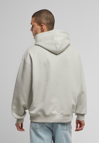 Lys grå hættetrøje med en stor hætte, dropped skuldre og ribbede manchetter. Blød tekstur, afslappet pasform og uden synlige logos. Denim jeans er iført.