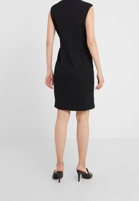 Robe noire sans manches avec une silhouette ajustée, dotée d'une couture à l'arrière et d'un ourlet à hauteur de genoux. Modèle portant des talons noirs à enfiler.