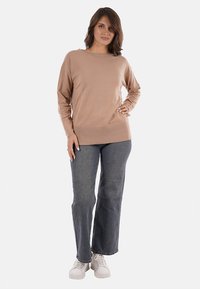 Donna in piedi con la mano sui fianchi, che indossa un top lungo beige, jeans blu e sneakers bianche su uno sfondo bianco.