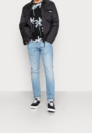 Sort pufferjakke med lynlås, sort t-shirt med palmetræsmønster, lyseblå jeans og sorte sneakers med hvide såler.