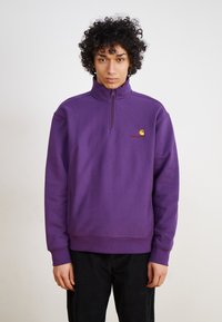 Lilla pullover sweatshirt med høj krave og kvart lynlås. Har et lille gult og rødt logo på brystet og ribbede ærmer.