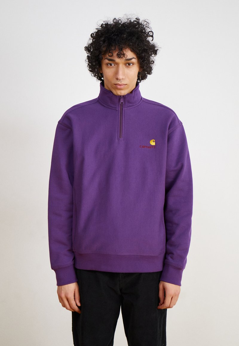 Lilla pullover sweatshirt med høj krave og kvart lynlås. Har et lille gult og rødt logo på brystet og ribbede ærmer.