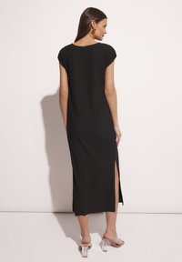 Czarna maxi sukienka o prążkowanej fakturze, z rękawami typu cap, bocznym rozcięciem i okrągłym dekoltem, zestawiona z przezroczystymi sandałami na obcasie.