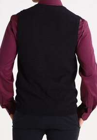 Veste noire tricotée avec un ourlet côtelé, portée sur une chemise bordeaux à manches longues. Texture lisse, design classique, sans motifs ni éléments métalliques visibles.