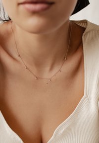 Różowe złoto naszyjnik z małymi, błyszczącymi akcentami wzdłuż delikatnego łańcuszka, noszony przez osobę w jasnej, prążkowanej bluzce.