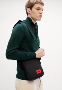 Schwarze Umhängetasche mit rotem HUGO-Logo-Patch, aus strapazierfähigem Gewebe gefertigt, mit einer eleganten rechteckigen Form und verstellbarem Tragegurt.