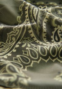 Gros plan sur un tissu vert olive avec des motifs beige paisley et floraux complexes, montrant les plis et les détails de la texture.