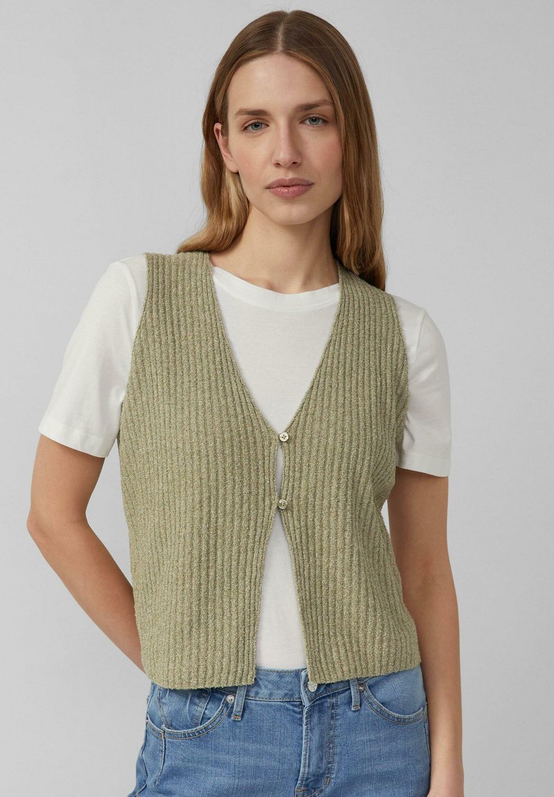 Frau trägt eine gerippte grüne Weste mit Knöpfen über einem weißen kurzärmligen Hemd und blauen Jeans, steht vor einem einfarbigen grauen Hintergrund.