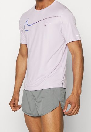 Mand iført en lyse lilla Nike løbetrøje og grå løbeshorts står med armene afslappet ned langs siderne.