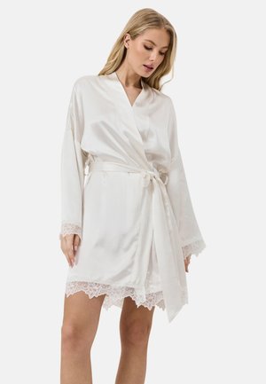 Vrouw draagt een witte satijnen badjas met kanten rand aan mouwen en zoom, gestrikt ter hoogte van de taille, staand tegen een effen achtergrond.