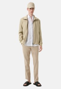 Beige lichte jas, witte linnen shirt, beige broek, zwarte sandaal, en een beige pet. Kenmerken zijn een minimalistisch ontwerp en een relaxte pasvorm.