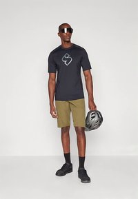 Μαύρο κοντομάνικο μπλουζάκι με λογότυπο, χακί σορτσάκι, μαύρες κάλτσες και μαύρα παπούτσια. Το μοντέλο κρατάει ένα γκρίζο κράνος, στέκοντας σε λευκό φόντο.