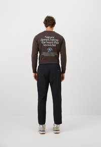 Brown long-sleeve shirt with white text on the back, paired with black pants and beige athletic shoes. Smooth fabric and relaxed fit.