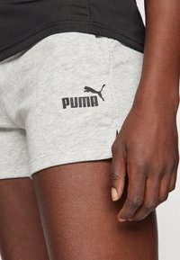 Harmaat puuvillashortsit, joissa on musta Puma-logo. Pitää sivuhalkiot hemmoissa ja teksturoitu musta paita päällä. Sileä kangaspinta.