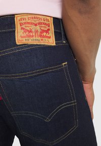 Tmavě modré džíny z denimu s žlutým prošíváním, klasickým designem pěti kapes a hnědou etiketou s červeným textem na zadním pasu.