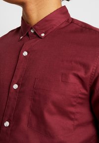 Detailní záběr na osobu v button-up vínové košili s bílými knoflíky a jednou náprsní kapsou, límec zapnutý knoflíky.