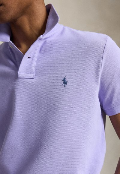 Hombre que lleva una camiseta polo de color púrpura claro con cuello levantado y un pequeño logo bordado de un jugador de polo en el pecho izquierdo.
